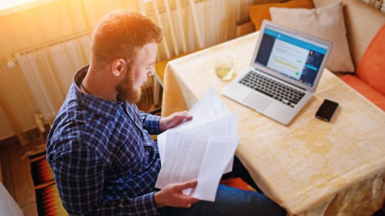 Ein Mann sitzt in seiner Wohnung am Laptop und studiert ausgedruckte Unterlagen