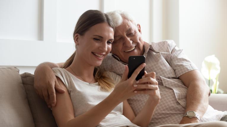 Eine Tochter sitzt mit ihrem Vater auf dem Sofa und sie blicken gemeinsam auf ein Smartphone.