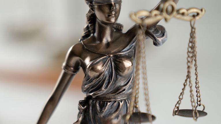 Eine Skulptur der Justitia auf einem Tisch, im Hintergrund der Hammer des Gerichts