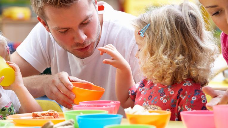Ein Mann sitzt mit Kindern an einem Kindertisch mit bunten Schüsseln beim Essen 