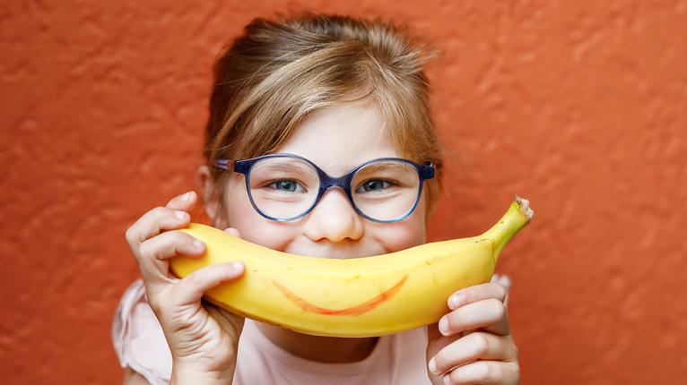 Ein Kind hält sich eine Banane mit aufgemaltem Lächeln vor den Mund