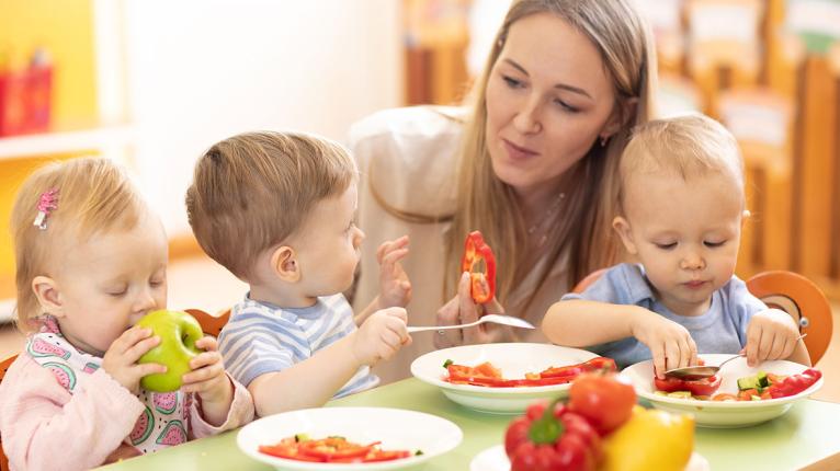 Eine Betreuerin sitzt mit drei Kleinkindern am Esstisch, auf dem Teller mit Gemüse stehen